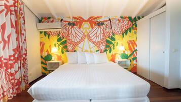 Habitación de hotel con una cama grande, dos mesitas de noche con lámparas, una pared con un mural de mariposas de colores y un ventilador de techo.