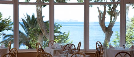 Un comedor con mesas dispuestas para una comida, ventanas amplias con vistas al mar y a los árboles.