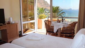 Habitación de hotel con cama, escritorio con cafetera, mesa con sillas y vista a la playa y al océano.