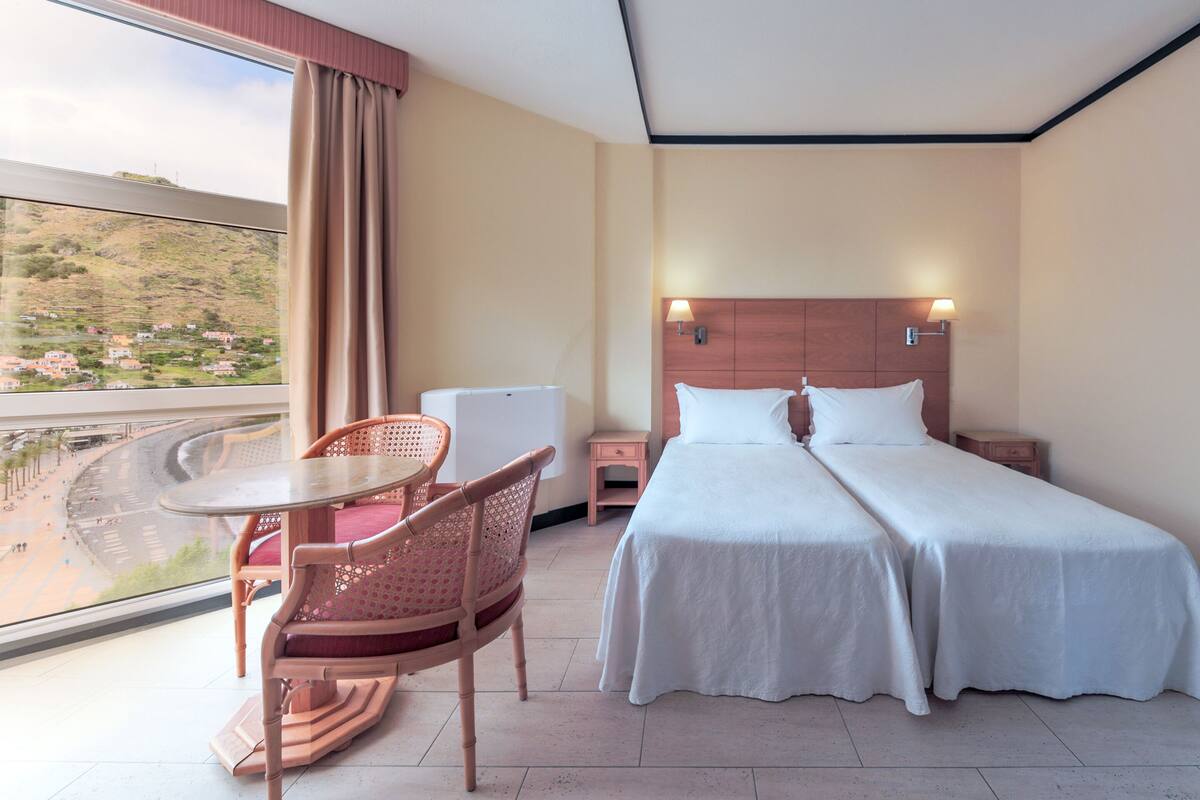 Ein Hotelzimmer mit Bett, Nachttischen, einem Sessel, einem kleinen Tisch und Blick auf Strand und Häuser durch das Fenster.