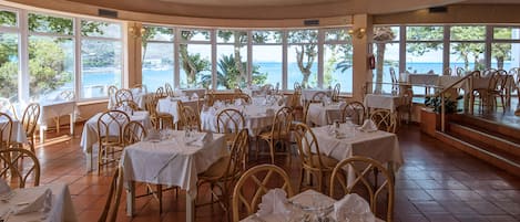 Um restaurante com várias mesas e cadeiras, janelas amplas com vista para o mar e teto de madeira.