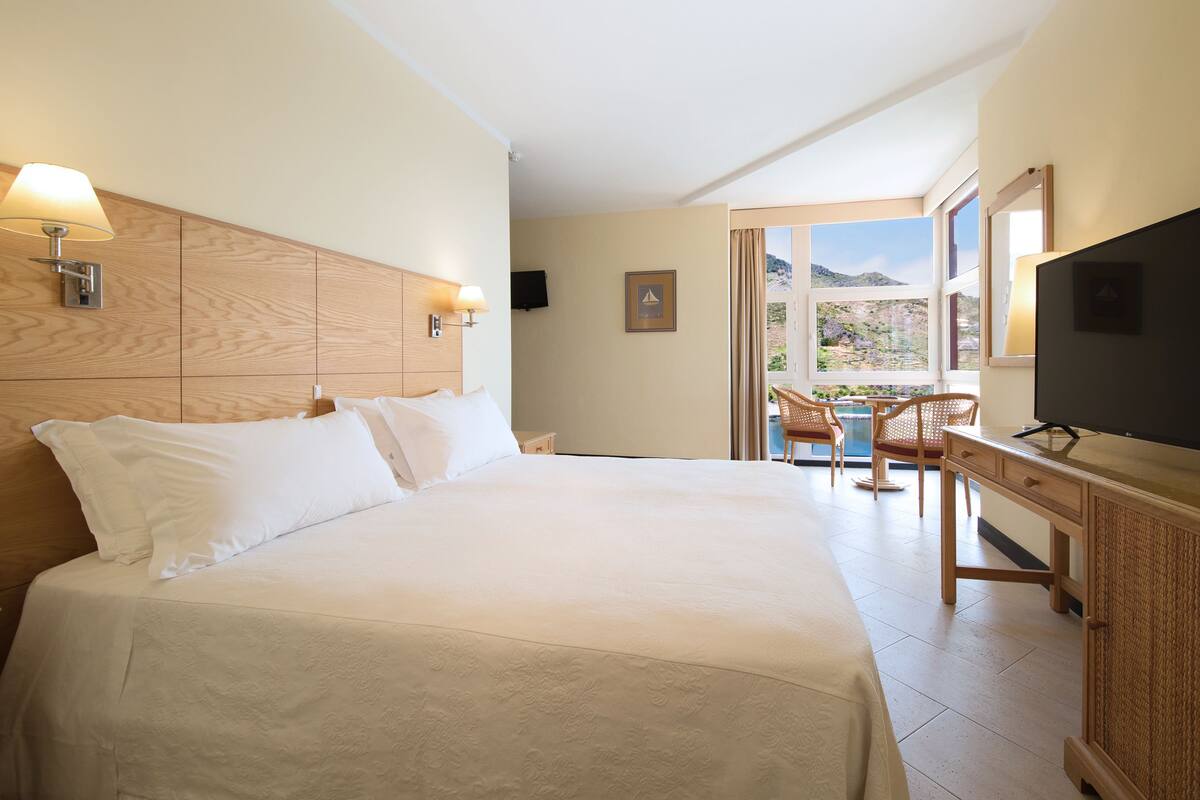 Ein Hotelzimmer mit einem großen Bett, einem Schreibtisch, einem Sessel, einem Fernseher und Blick ins Freie.