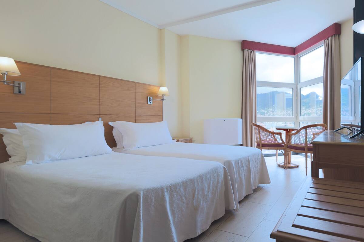 Ein Hotelzimmer mit zwei Betten, einem Schreibtisch, einem Stuhl und einem großen Fenster mit Blick auf die Stadt.