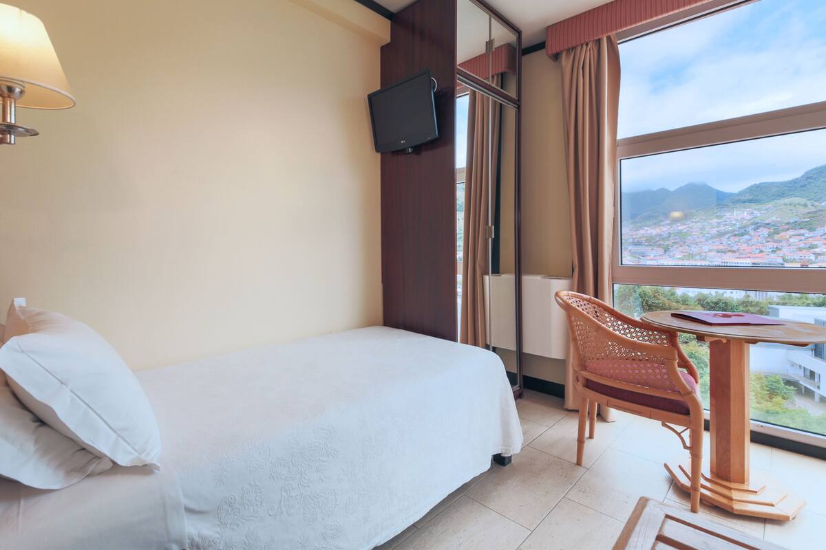 Ein Hotelzimmer mit einem Bett, einem an einer Holzkommode befestigten Fernseher, einem Tisch mit einem Stuhl und einem großen Fenster mit Blick auf die Stadt.
