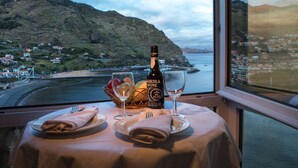 Una mesa con una botella de vino, copas y una cesta de fruta, con vistas a un pueblo costero y colinas.