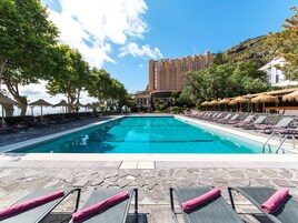 Una piscina con sillones de descanso y sombrillas, rodeada de árboles y un edificio al fondo.