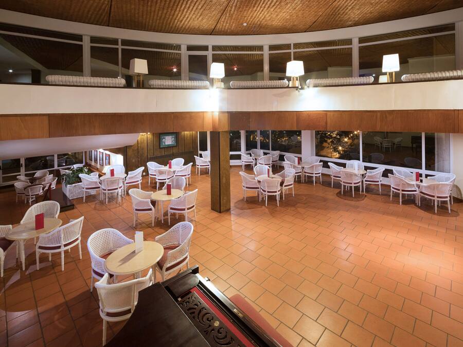A spacious indoor area with multiple white wicker tables and chairs, large windows, and a high ceiling with a woven pattern.