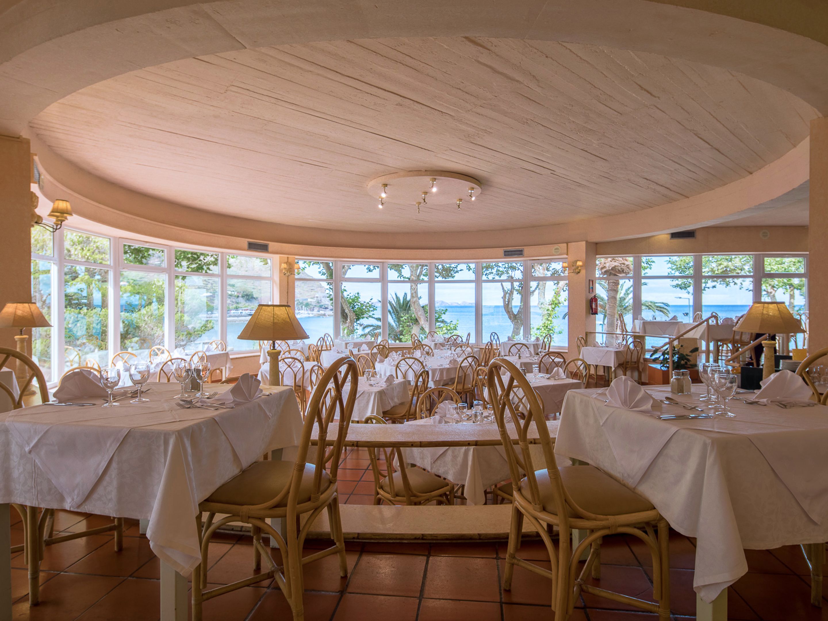En restaurang med runda bord och stolar av rotting, stora fönster med utsikt över havet och ett tak av trä.