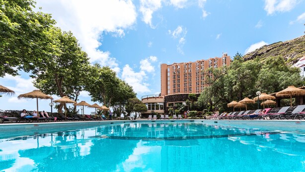 Una piscina con sillas de descanso y sombrillas, rodeada de árboles y un edificio alto.