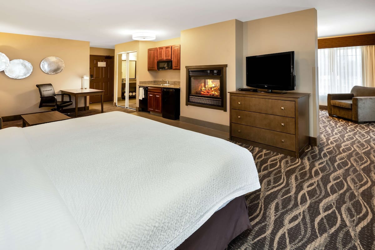 A hotel room with a large bed, a television, a fireplace, a desk with a chair, and a kitchenette.