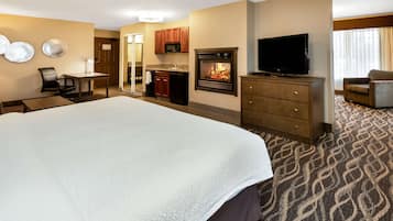 A hotel room with a large bed, a television, a fireplace, a desk with a chair, and a kitchenette.
