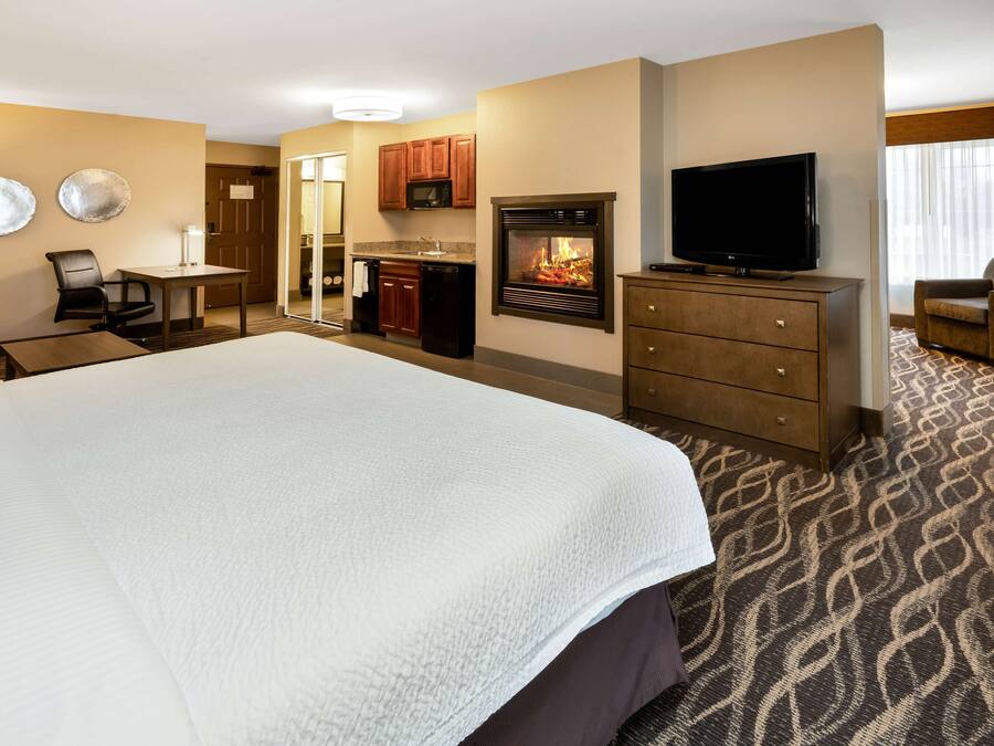 A hotel room with a large bed, a television, a fireplace, a desk with a chair, and a kitchenette.