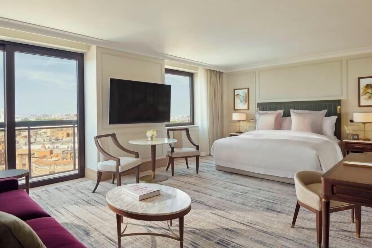A hotel room with a large bed, a TV, a small table with chairs, and a view of the city through large windows.