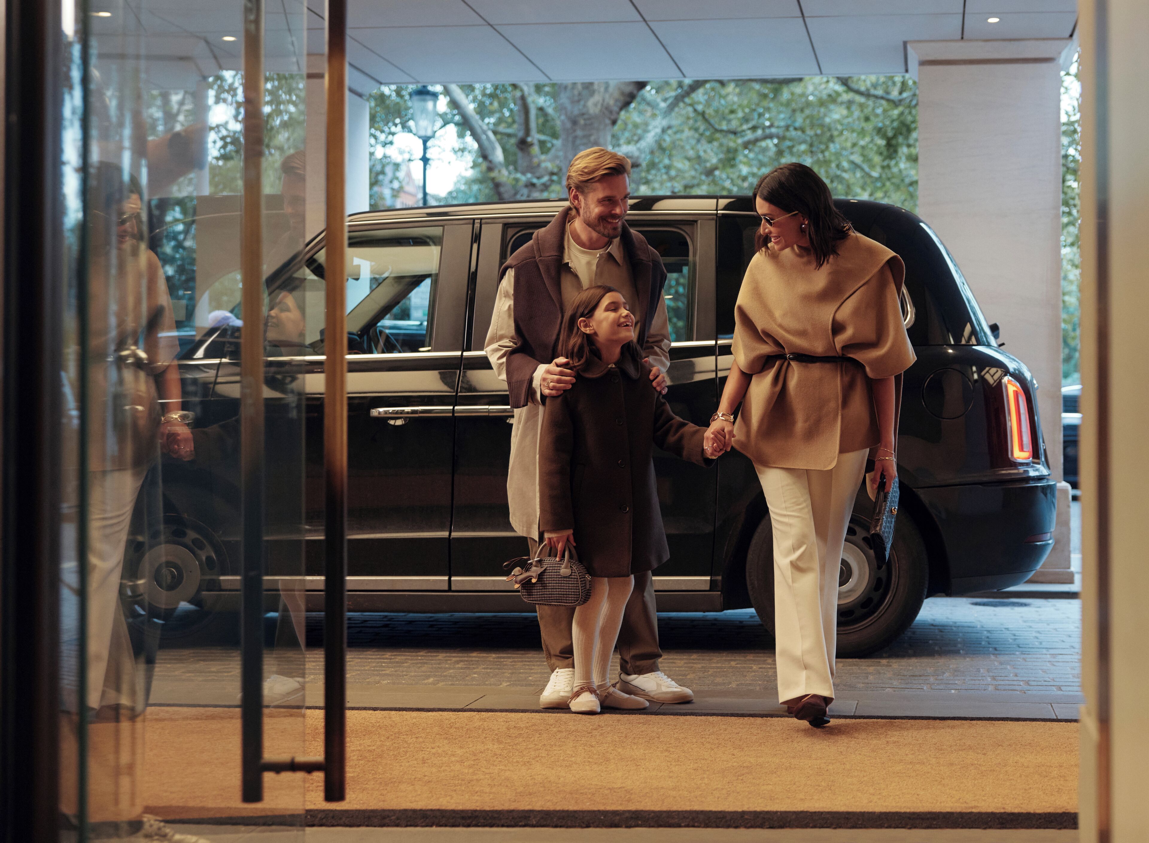 A family holding hands in front of a black SUV, with a glass door reflecting a person inside.