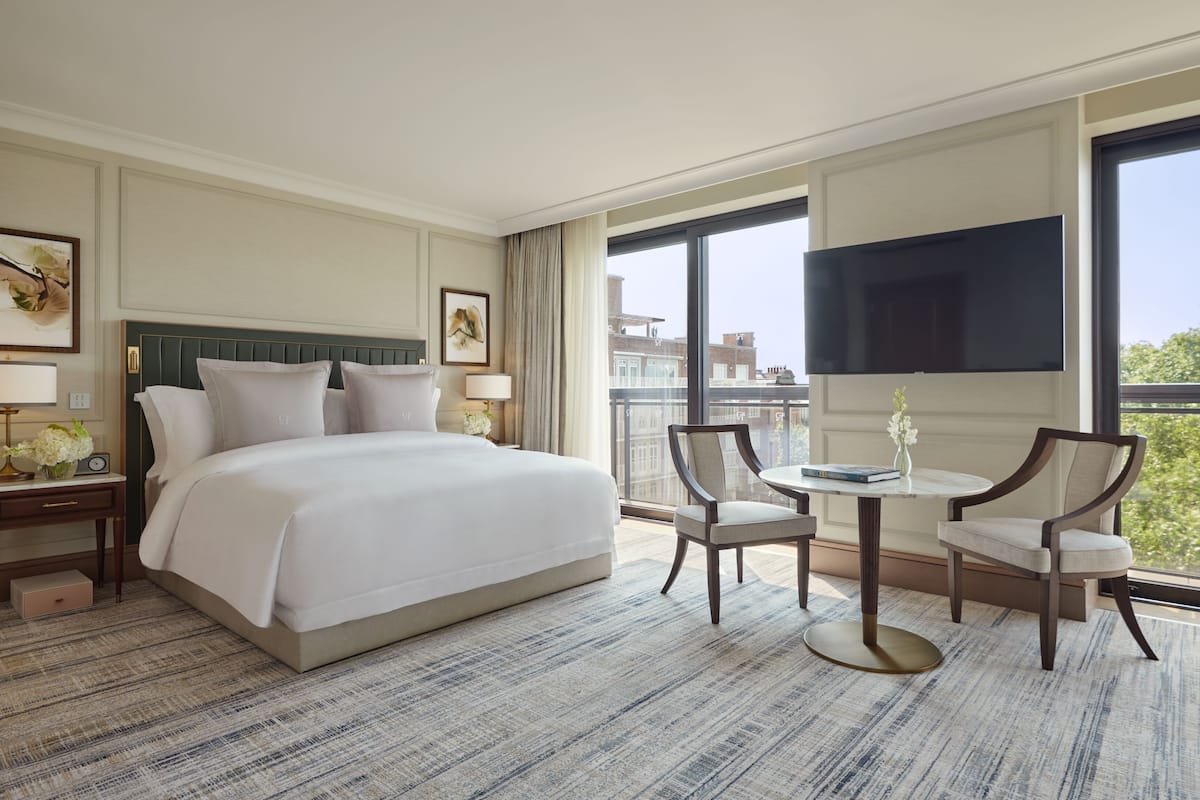 Habitación de hotel con una cama grande, dos mesitas de noche con lámparas, una pequeña mesa con sillas, un televisor montado en la pared y un balcón con vistas.