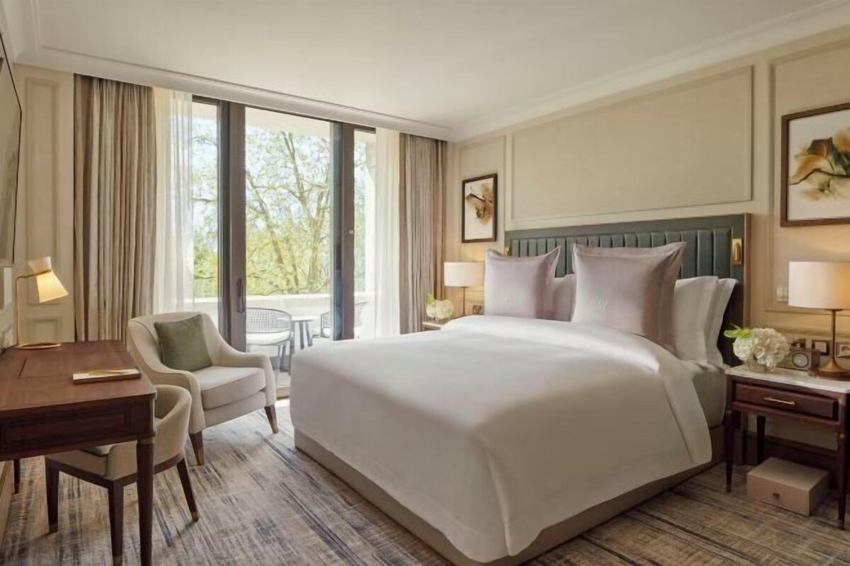 A hotel room with a large bed, a desk, a chair, and a view of trees through the window.