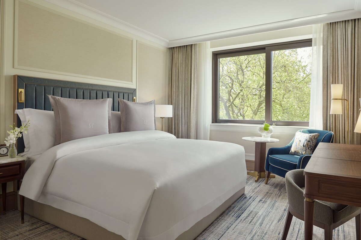 A hotel room with a large bed, a desk, a chair, a small table, and a window with a view of trees.