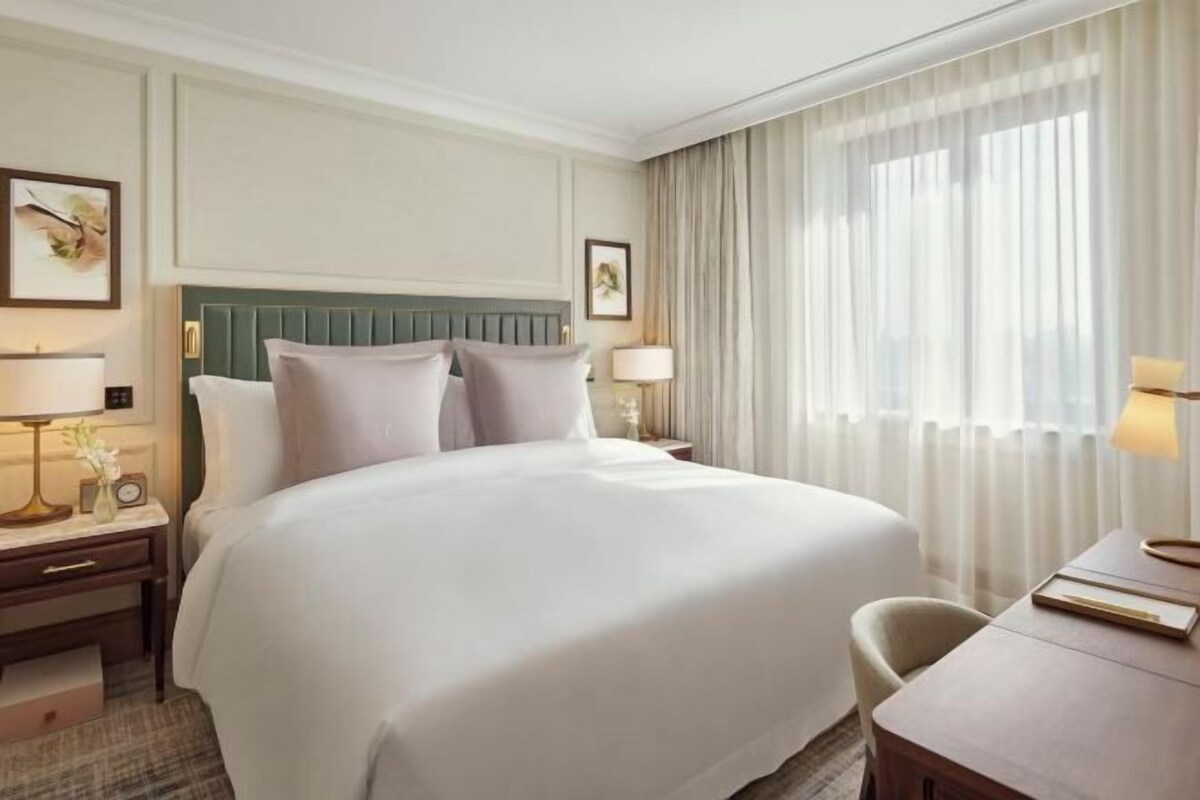 A hotel room with a large bed, a desk, a chair, a nightstand, and a window with curtains.