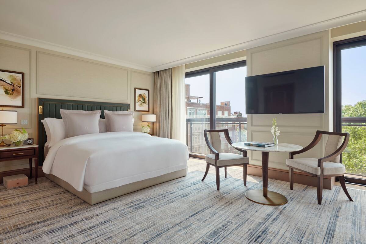 Habitación de hotel con una cama grande, dos mesitas de noche con lámparas, una pequeña mesa con sillas, un televisor montado en la pared y un balcón con vistas a edificios.