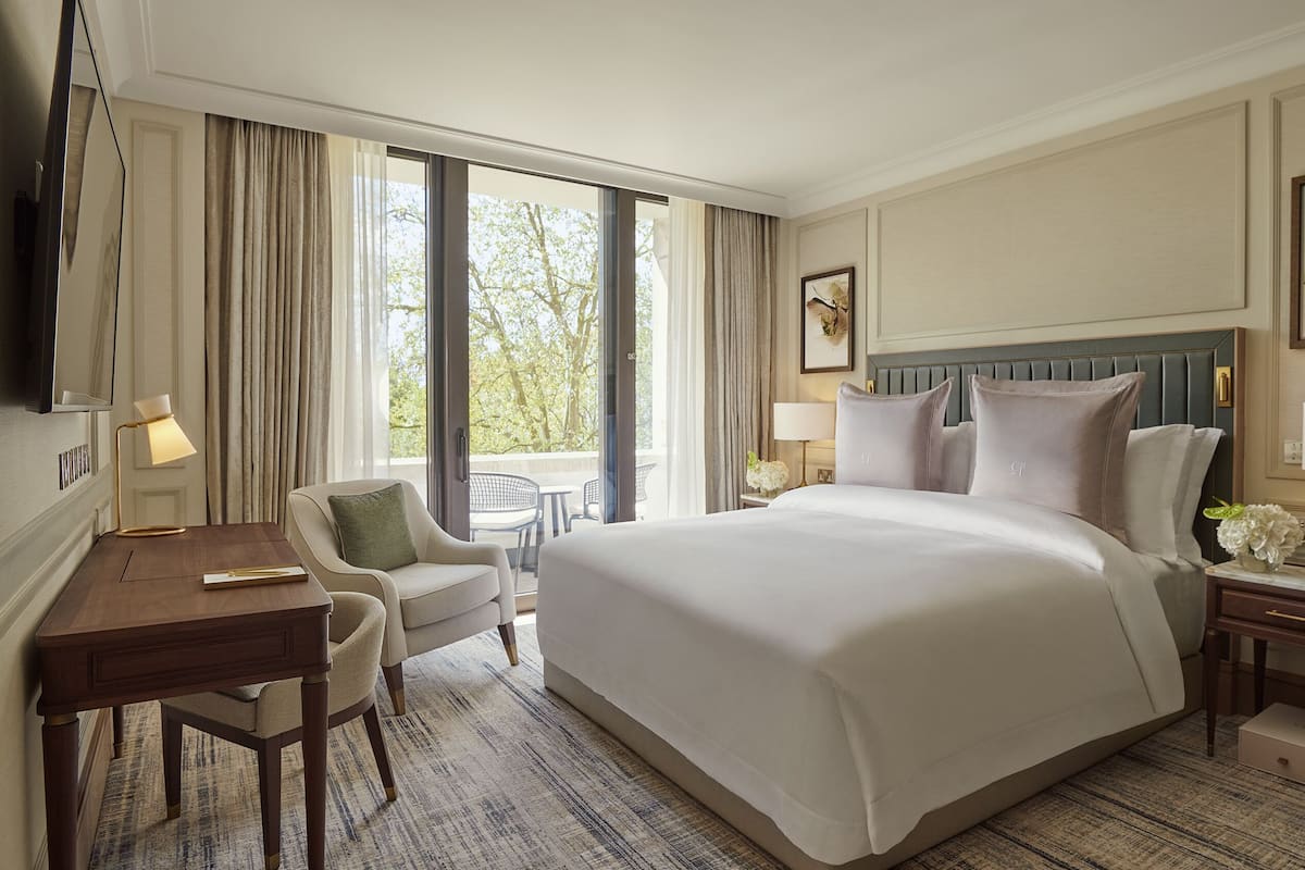 Habitación de hotel con una cama grande, un escritorio con silla, una mesita y vistas a árboles por la ventana.