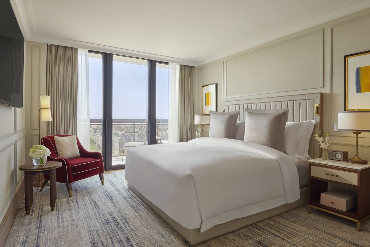 Habitación de hotel con una cama grande, un sillón rojo, una mesita con un jarrón de flores, una mesita de noche con lámpara y vistas a la ciudad a través de amplios ventanales.
