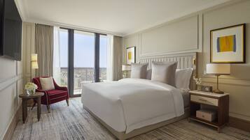 A hotel room with a large bed, a red armchair, a small table with a vase of flowers, a nightstand with a lamp, and a view of the city through large windows.