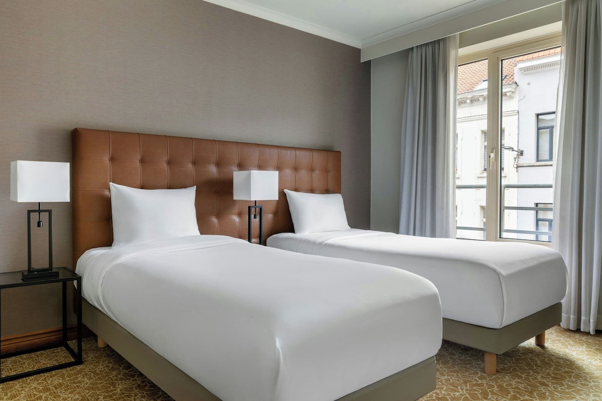 A hotel room with two beds, a brown headboard, white bedding, a nightstand with a lamp, and a view of the outside through a window.