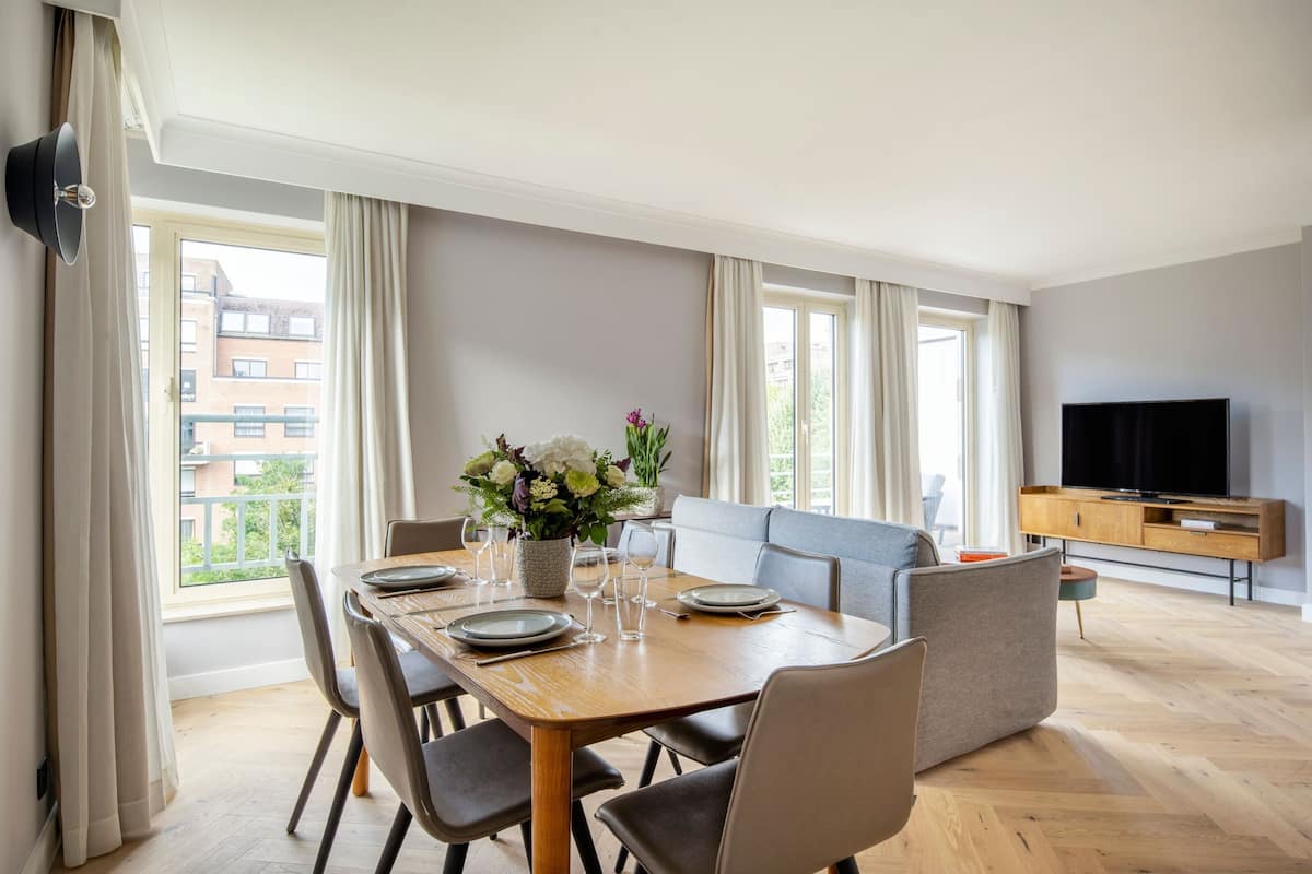 A modern living room with a dining area, large windows, a flat-screen TV, and a sofa.