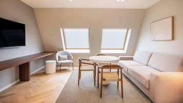A modern living room with a sofa, a round table, and two chairs. There is a flat-screen TV mounted on the wall, and two windows with blinds.