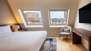 A modern hotel room with a large bed, a desk, a chair, and two windows offering views of the cityscape. - Marriott Executive Apartments Brussels, European Quarter (Brussels)