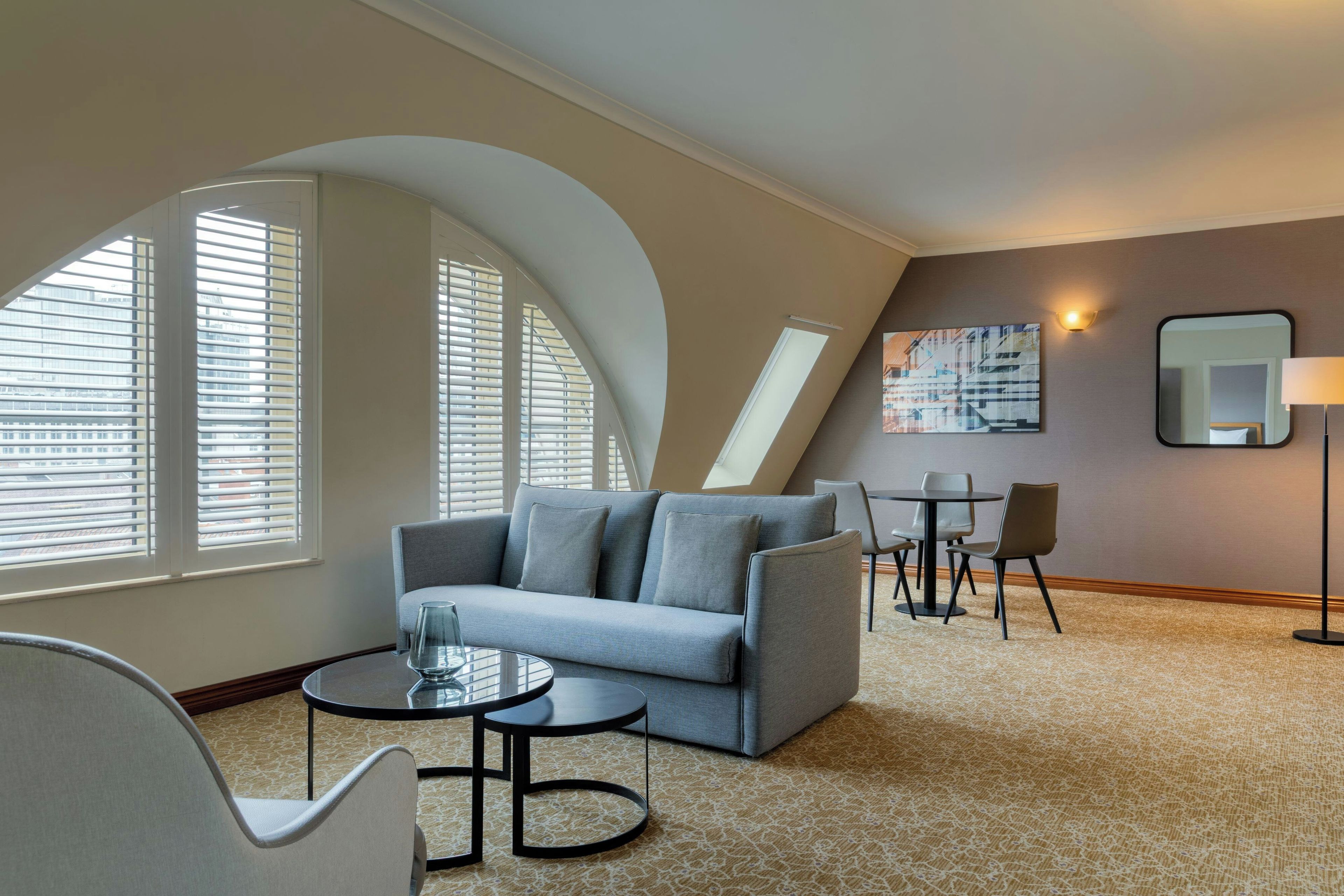 Une chambre d’hôtel moderne avec un plafond incurvé, un canapé gris, une petite table ronde, une chaise et un tableau au mur.