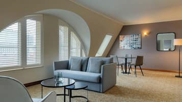 Une chambre d’hôtel moderne avec un plafond incurvé, un canapé gris, une petite table ronde, une chaise et un tableau au mur.