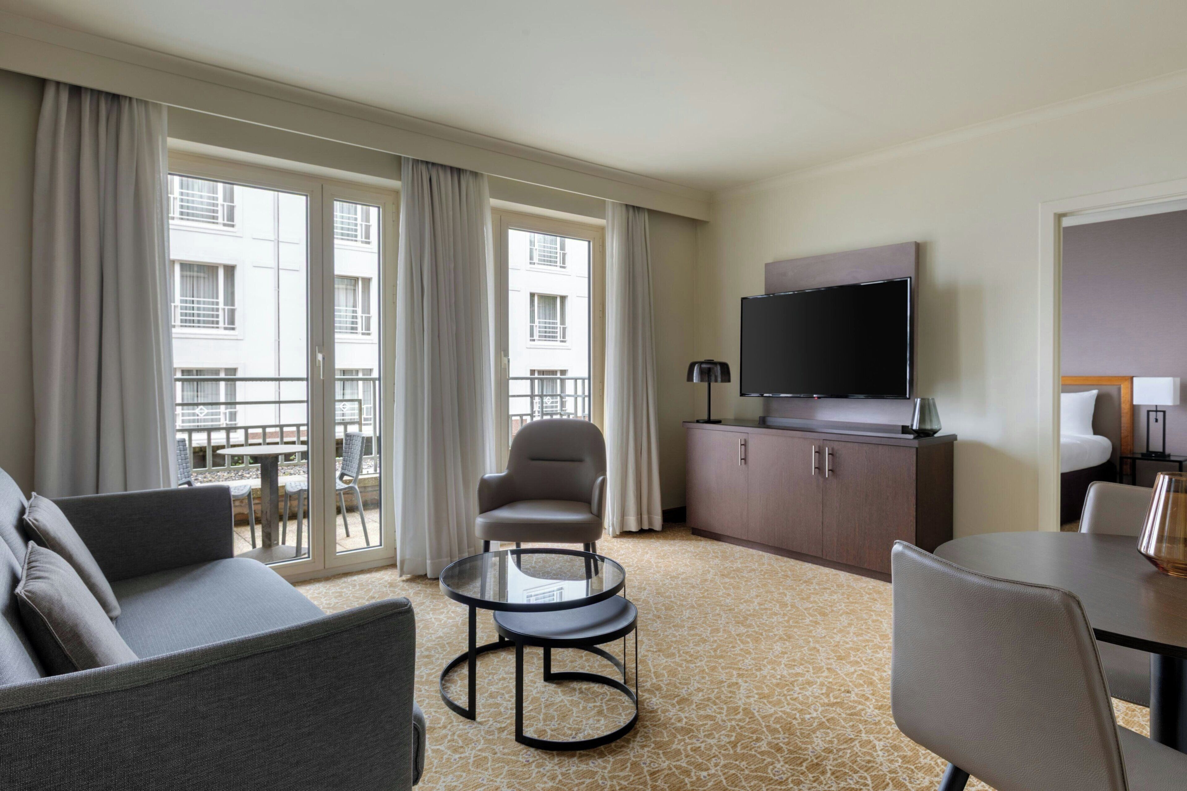 A hotel room with a sofa, armchair, small round table, a TV on a cabinet, and a balcony with curtains.
