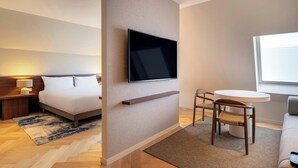 A modern hotel room with a bed, a flat-screen TV mounted on the wall, a small round table with a chair, and a window providing natural light. - Marriott Executive Apartments Brussels, European Quarter (Brussels)