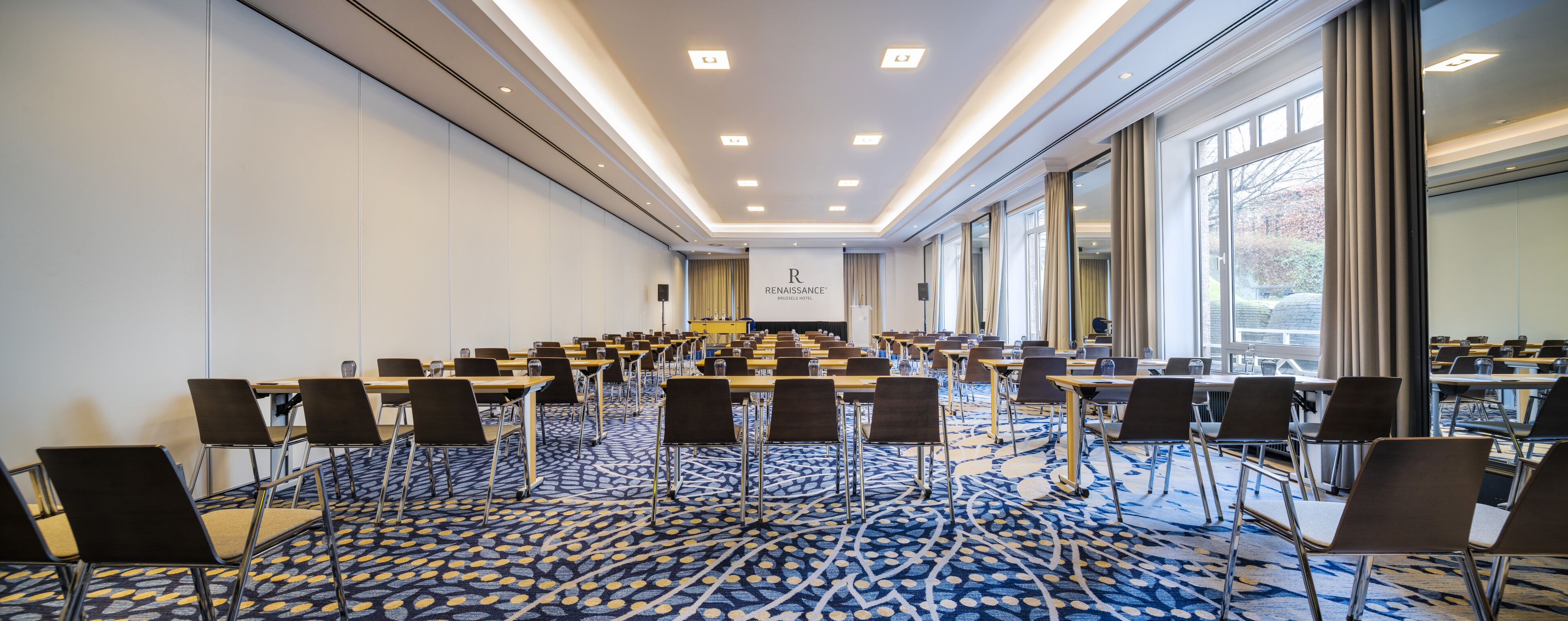 A conference room with rows of tables and chairs, large windows, and a patterned carpet.