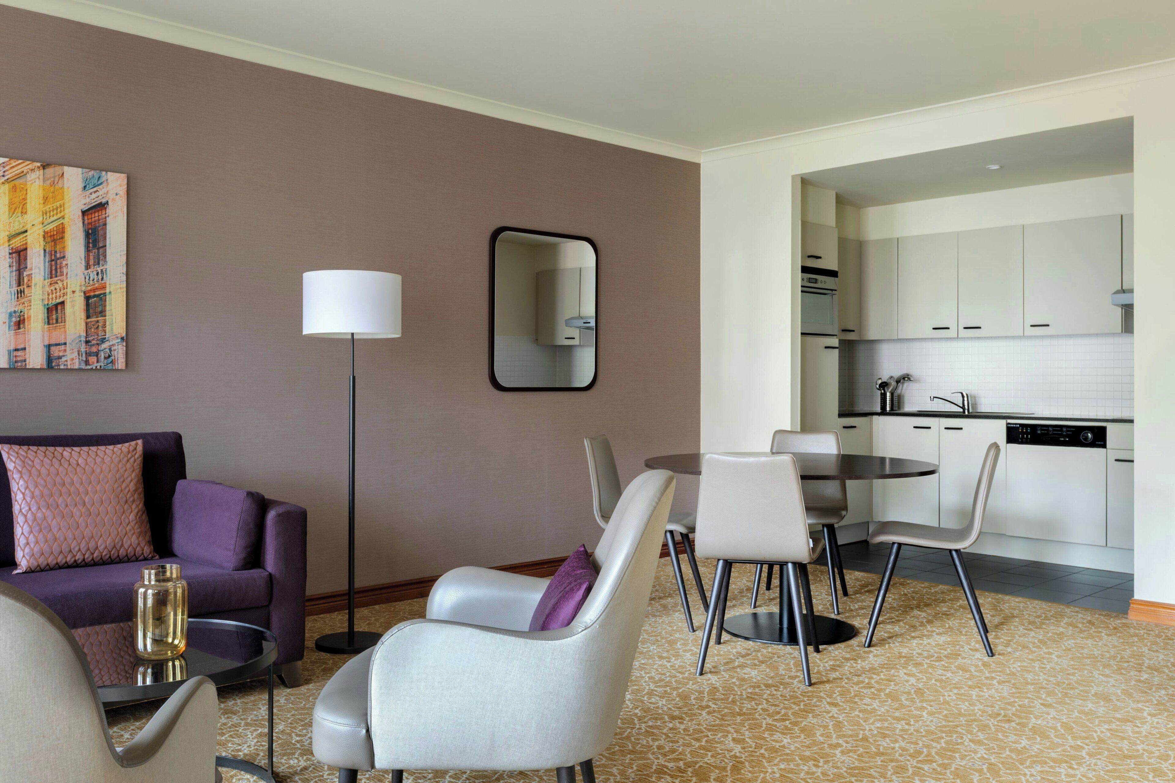 A modern hotel room with a sofa, chairs, a dining table, a floor lamp, a mirror, and a kitchen area.