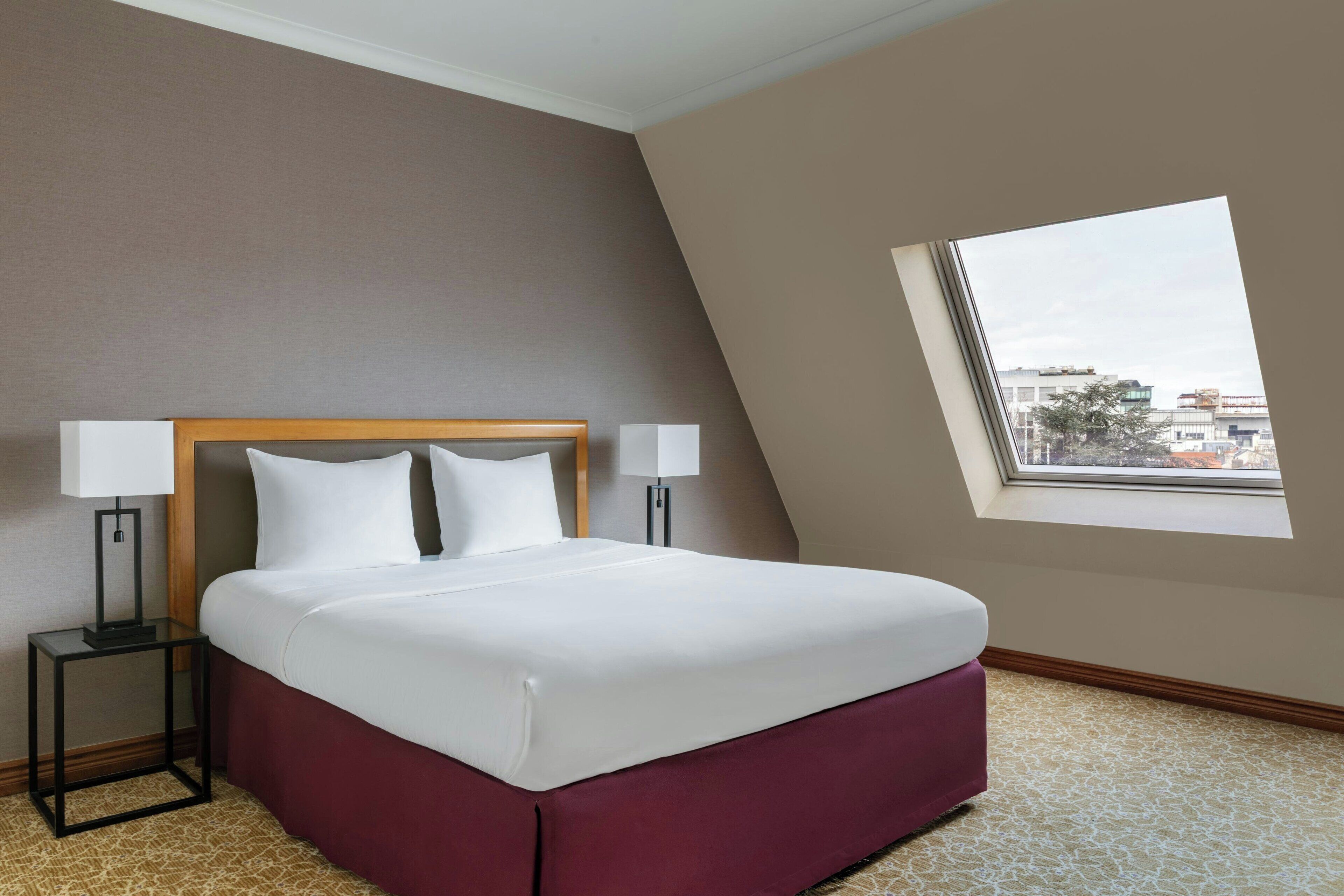 Une chambre d’hôtel avec un lit, deux lampes de chevet, une fenêtre donnant sur des immeubles et un sol recouvert de moquette.