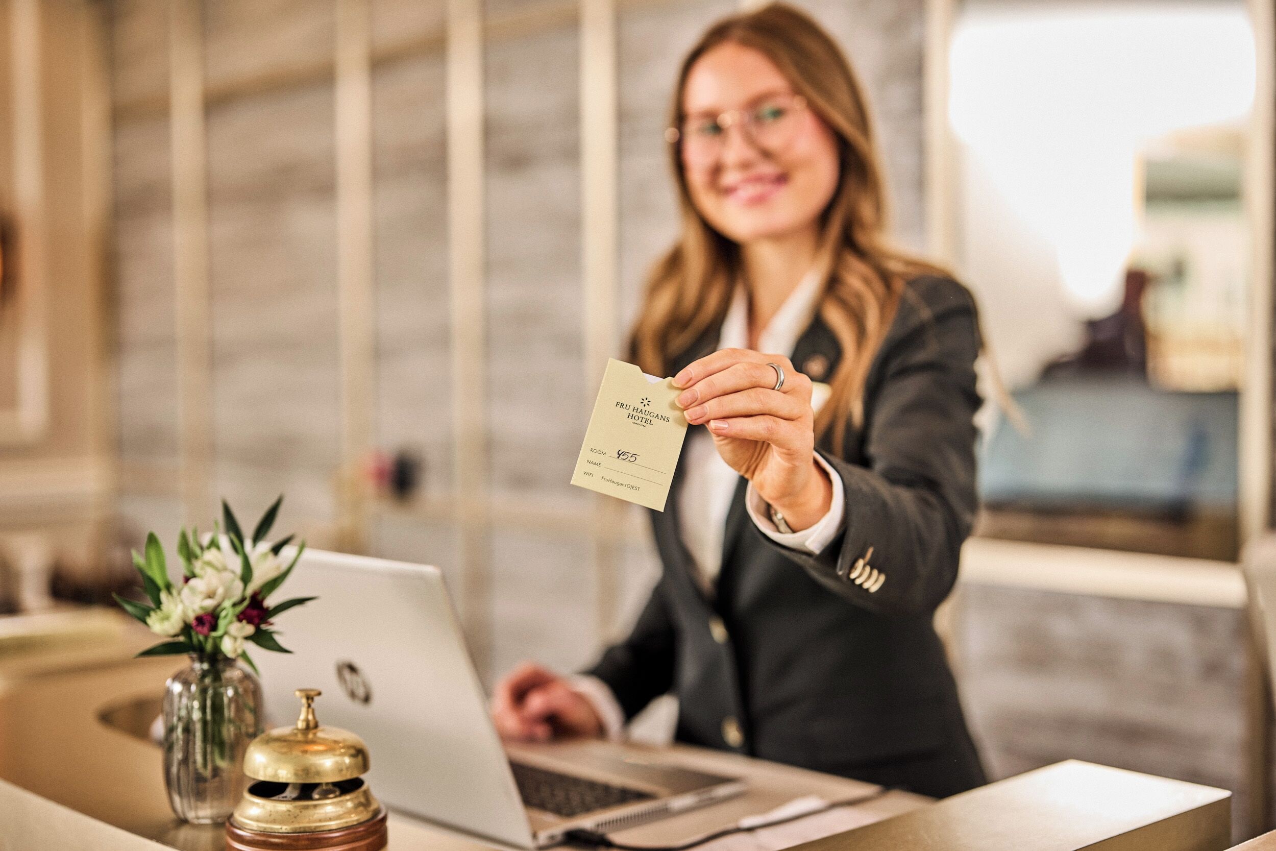 A person holding a card at a reception desk with a laptop, a vase with flowers, and a bell.