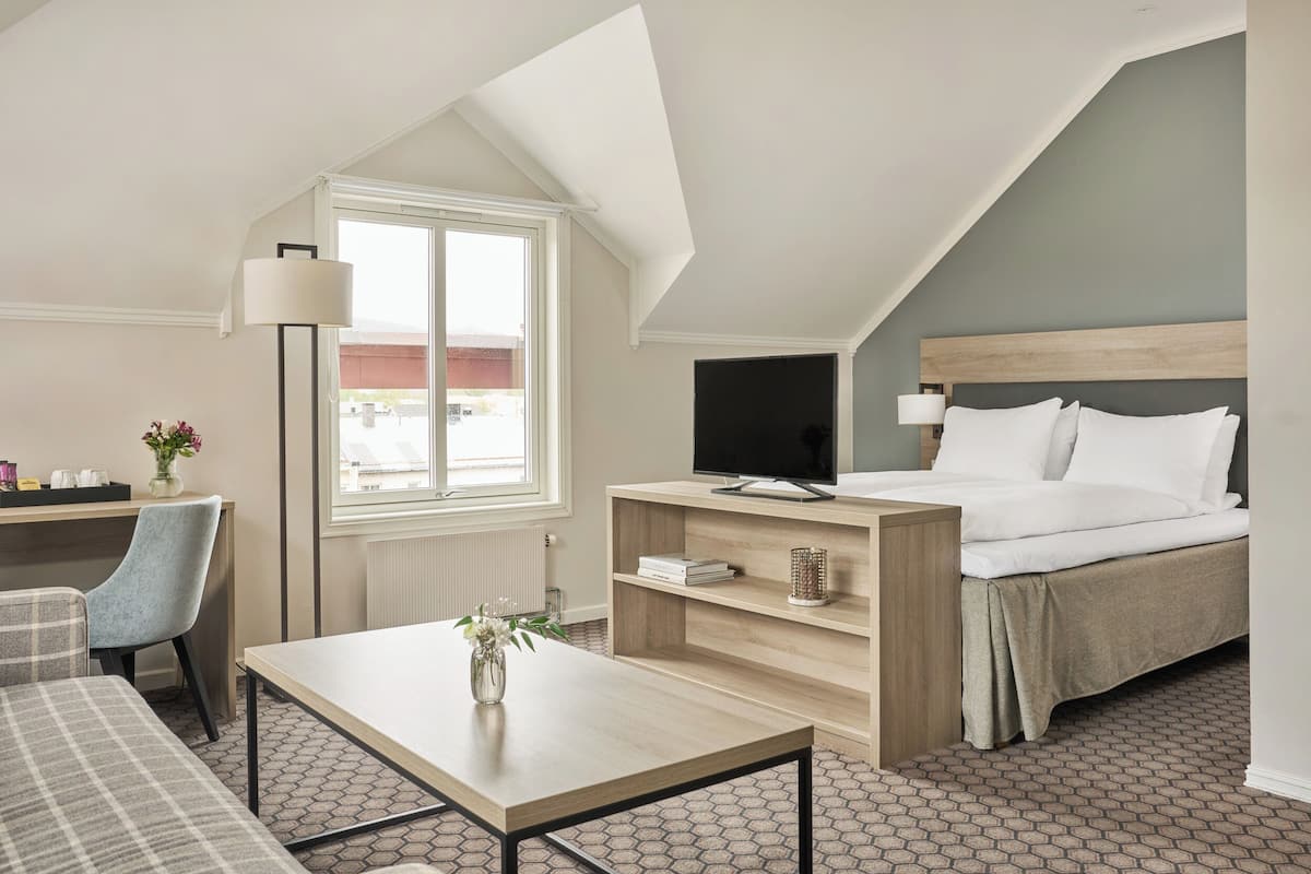 A hotel room with a bed, a desk with a computer, a chair, a coffee table, and a window with a view of the outside.