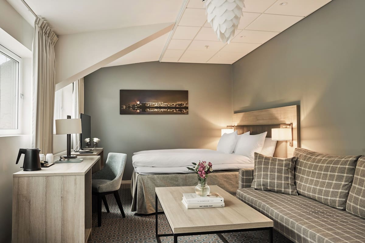 A modern hotel room with a bed, a sofa, a desk, and a coffee table. There is a cityscape view on the wall, a window with curtains, and a lamp on the desk.