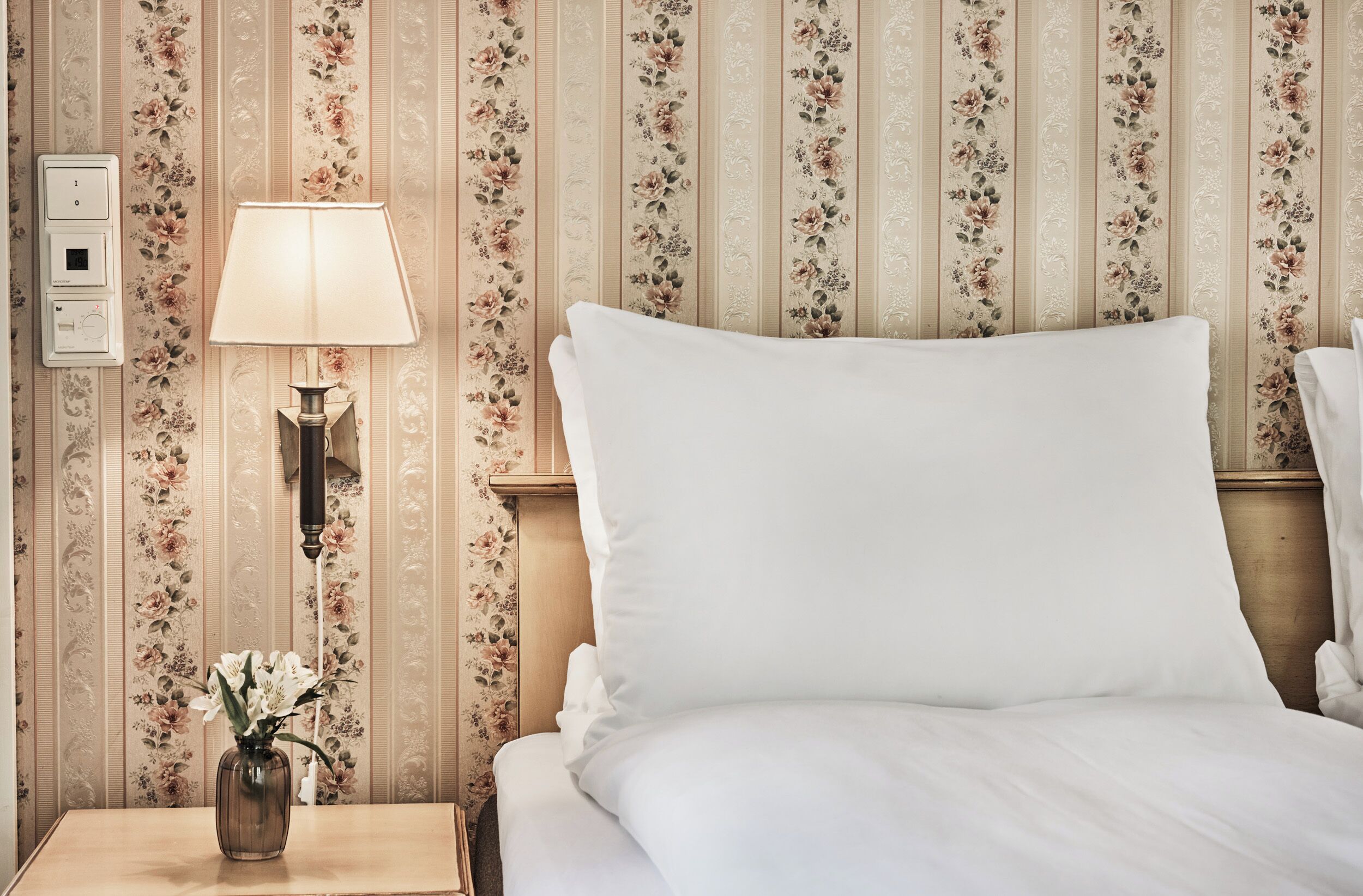 A hotel room with a bed, a nightstand with a lamp, and a vase with flowers.