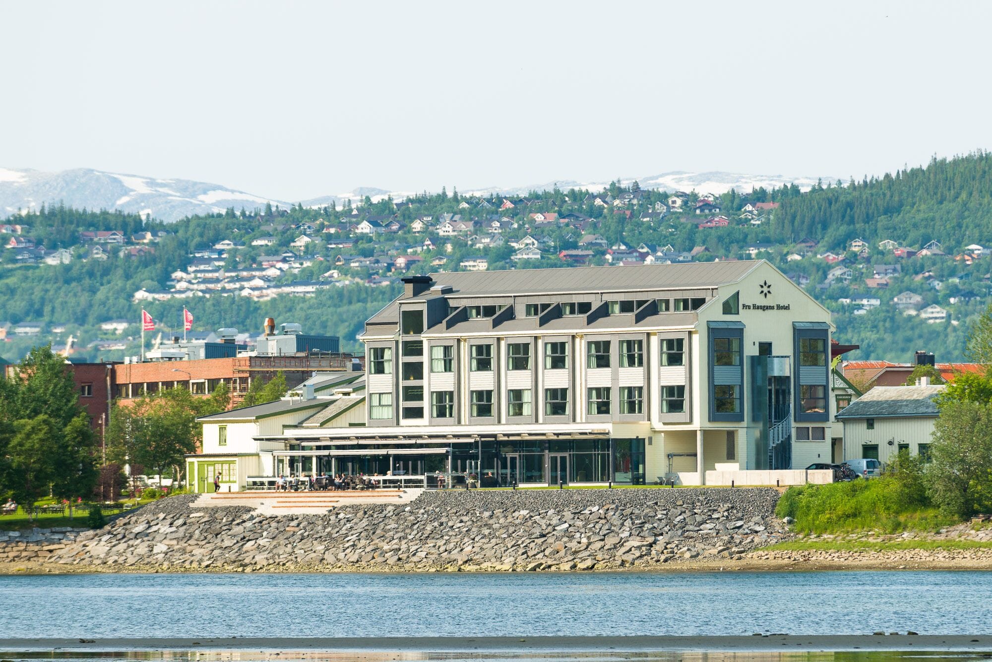 A modern hotel with a flat roof and large windows, situated by a lake with a rocky shoreline.