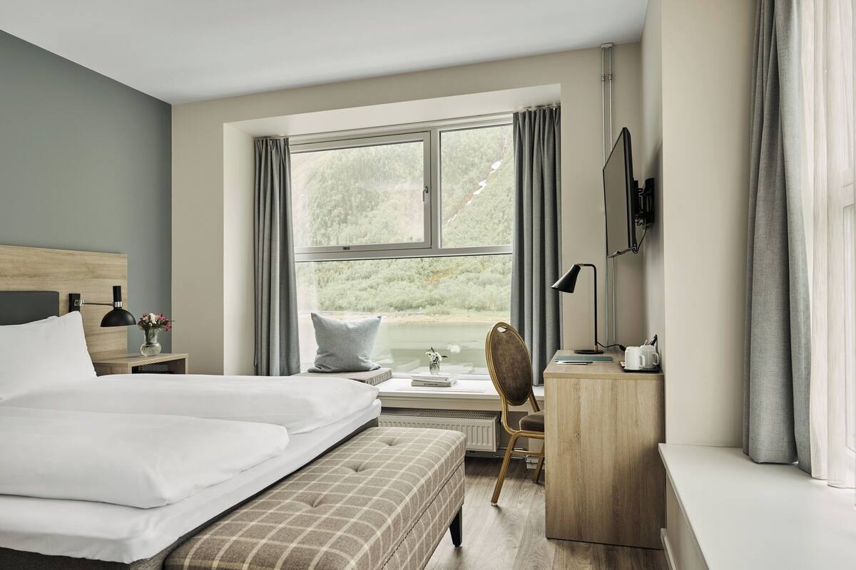 A hotel room with a large bed, a desk with a chair, a window with curtains, a bedside table with a lamp, and a TV mounted on the wall.