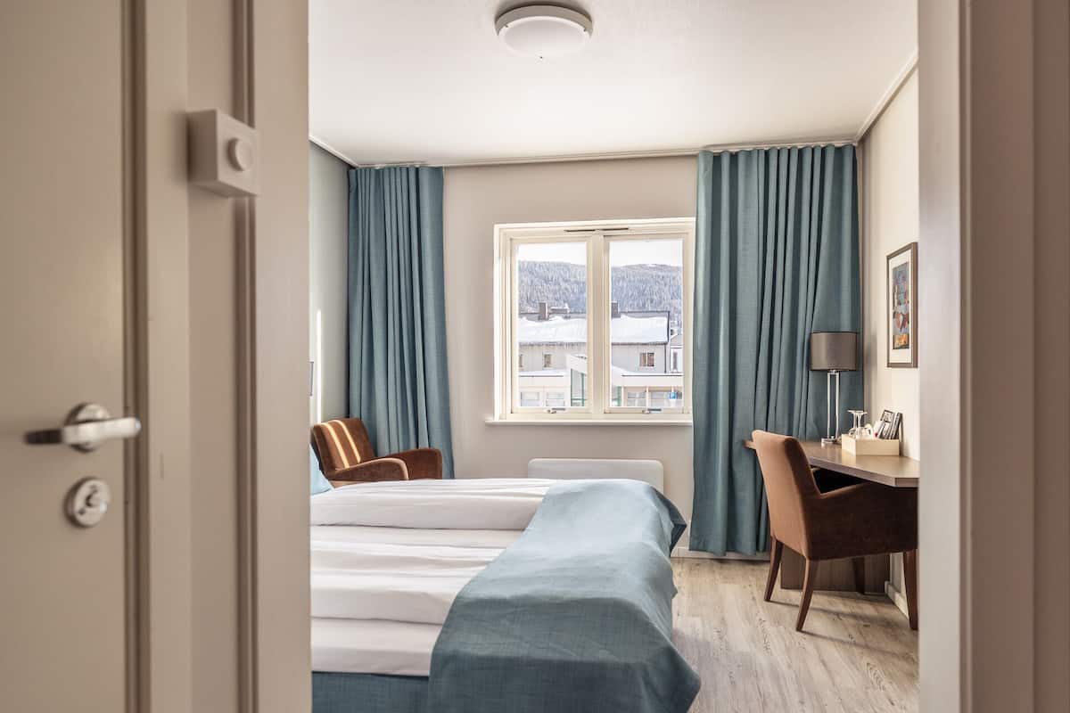 A hotel room with a bed, a desk, a chair, a window with a view of buildings, and a door.