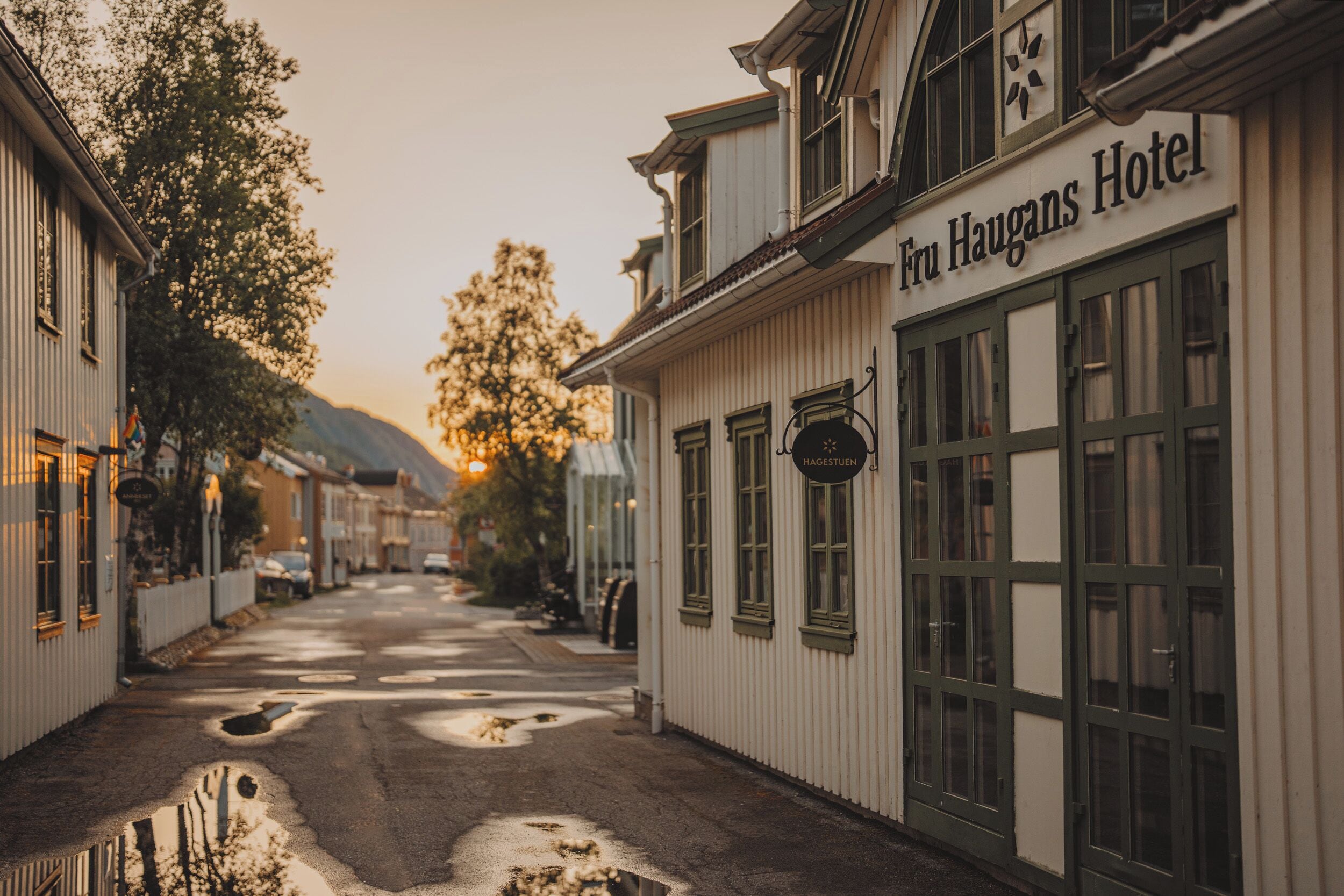 A street view of the Fru Haugans Hotel with a clear sign, a narrow road with puddles, and buildings on either side.