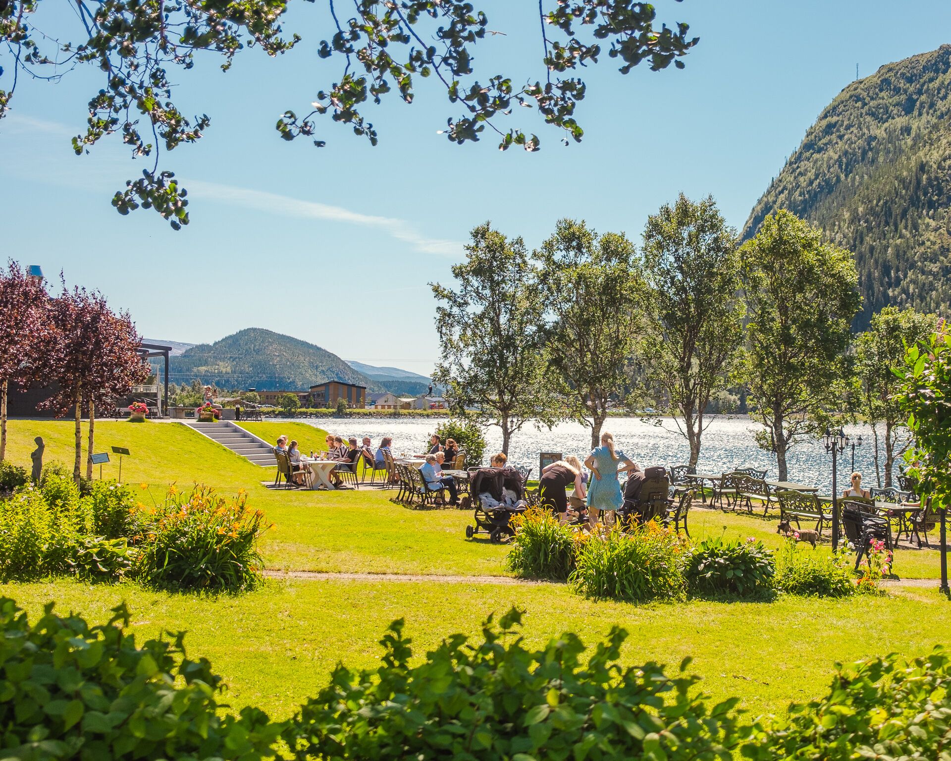 A lakeside dining area with outdoor seating, surrounded by lush greenery and mountains in the background.