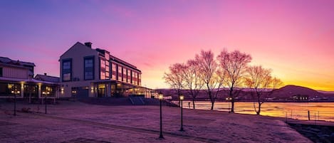 A building with a flat roof and large windows, situated near a waterfront with a sunset in the background.