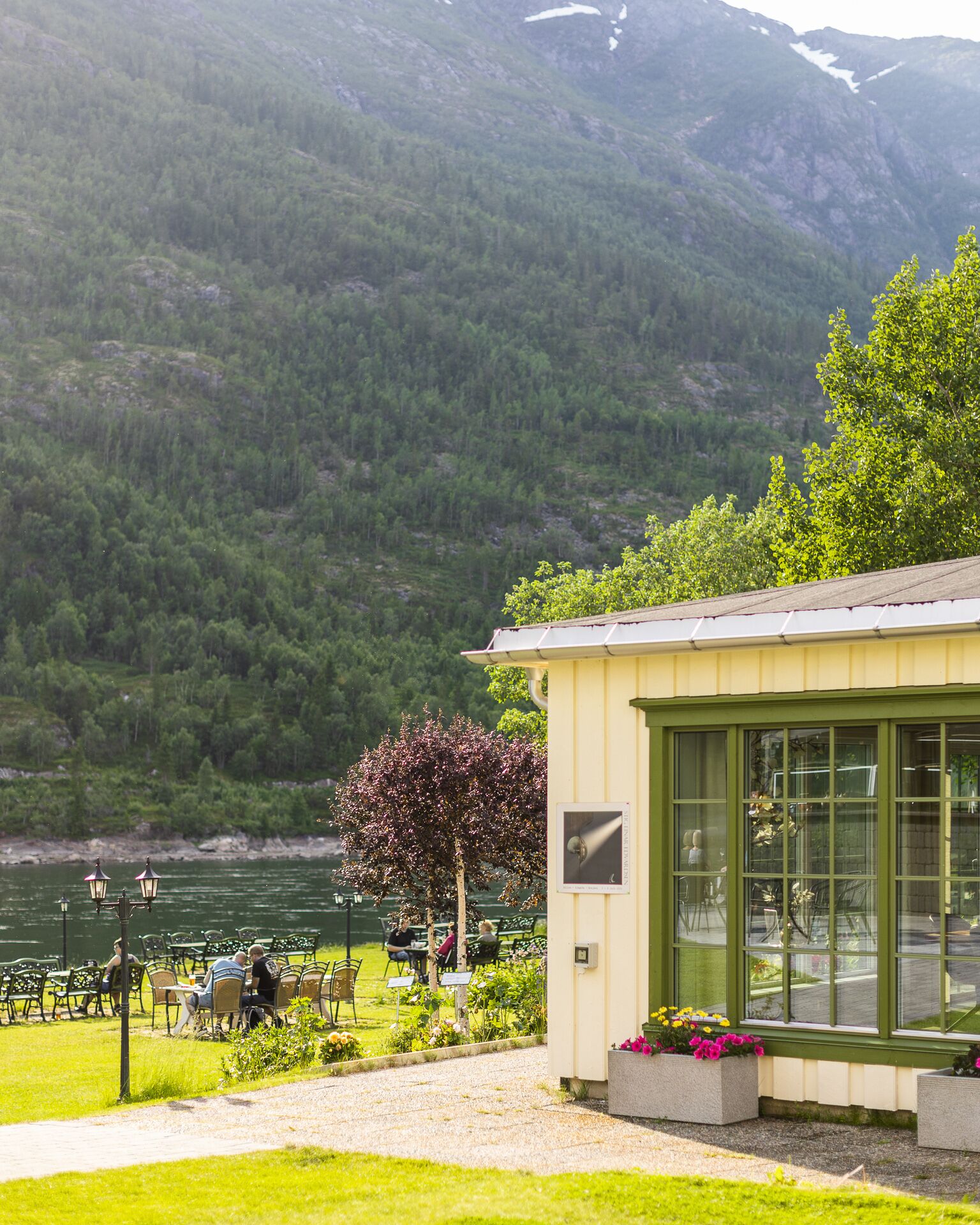 A lakeside building with a patio area, surrounded by greenery and mountains.