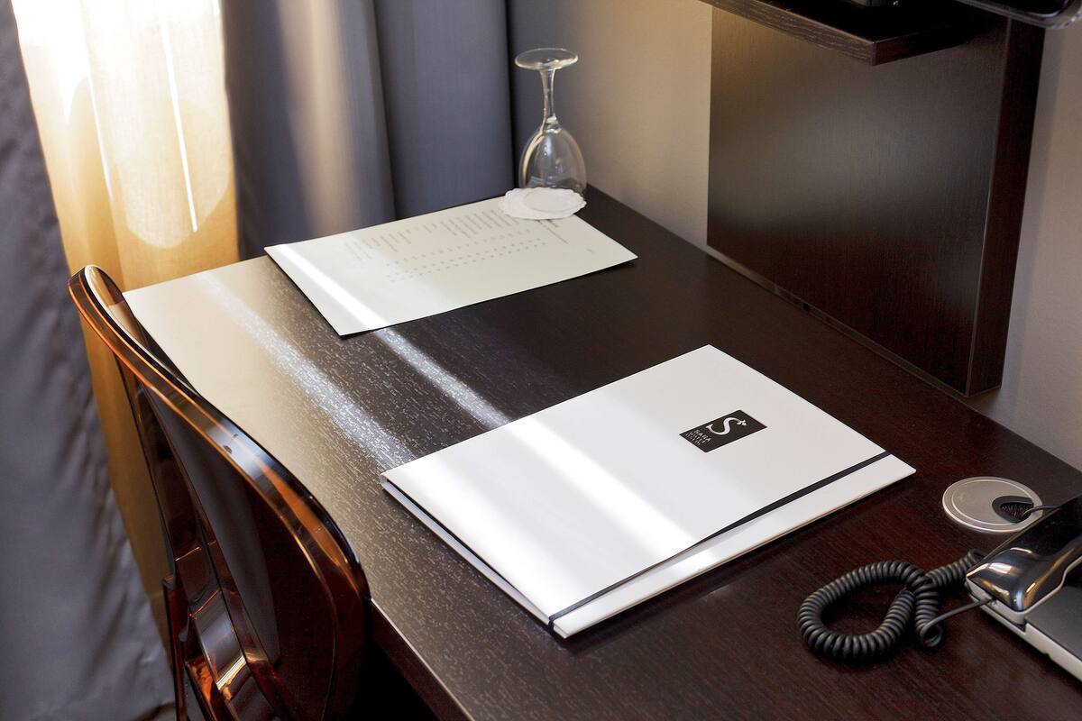 A hotel room desk with a laptop, a glass of water, a phone, and a menu.