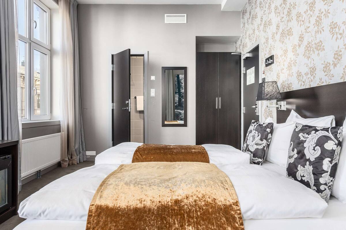 A hotel room with a large bed, a dresser, a window with curtains, a radiator, and a wall-mounted lamp.